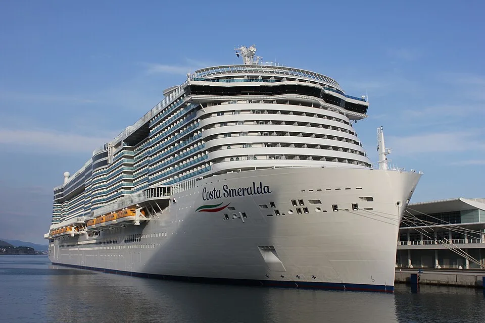 The massive white cruise ship Costa Smeralda is docked at a port terminal on calm blue water. The vessel features its name clearly printed on the side, yellow lifeboats tucked into its lower deck, and numerous towering tiers of passenger cabins under a bright, clear sky.