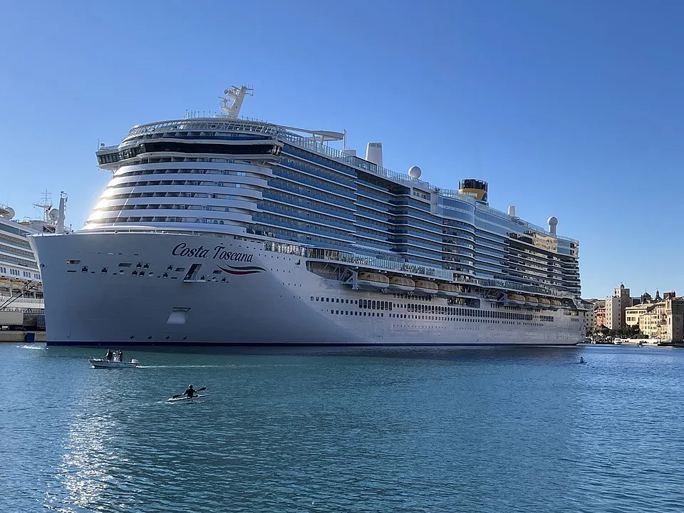 The massive cruise ship "Costa Toscana" sits in bright blue water under a clear sky. It has countless stacked balconies, a yellow funnel, and the ship name printed on its large bow. A small motorboat and a person in a kayak stir up the water in the foreground, with buildings on the right shore.