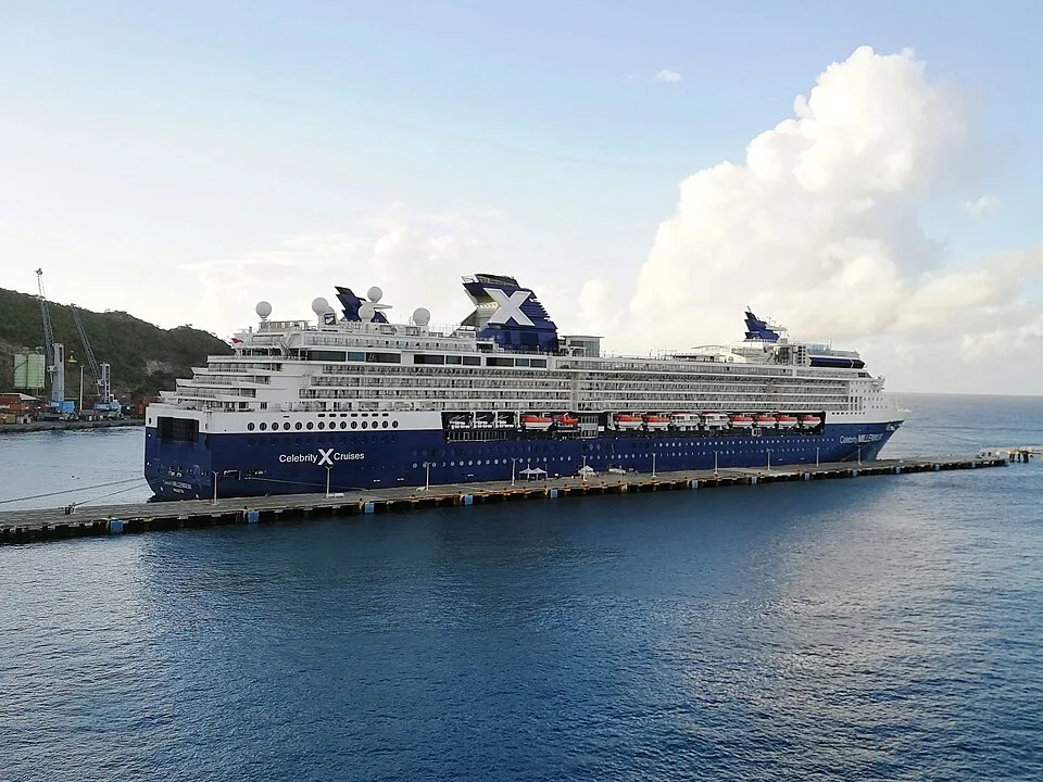 The Celebrity Millennium cruise ship docked at a long pier. The large white and dark blue ship features multiple cabin decks, orange lifeboats, and a prominent "X" logo on its funnel under a partly cloudy sky.