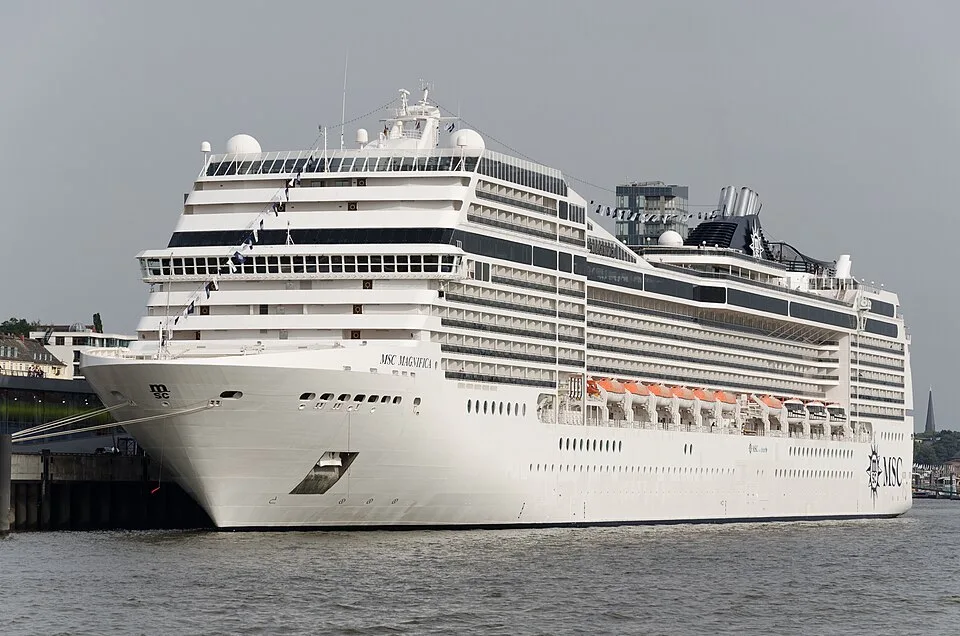 A large white cruise ship named "MSC MAGNIFICA" is docked along a pier on murky water under an overcast sky. The ship features tiered passenger decks, rows of balconies, orange lifeboats along the side, and a dark funnel toward the rear. The MSC logo is printed near the back of the hull.