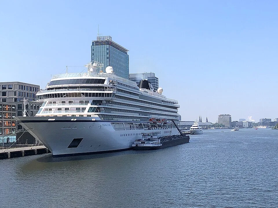 A large white Viking cruise ship, named "Viking Star," docked at a city port along a wide river. A small barge is tied to its side, with city buildings and a tall glass skyscraper visible in the background under a clear sky.