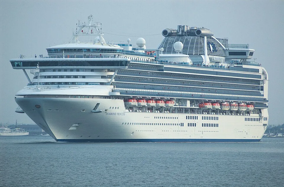 The Diamond Princess cruise ship sails on calm gray water. The massive white vessel features a wide, multi-level glass navigation bridge, numerous decks lined with windows, a row of orange lifeboats, and a unique blue-and-white funnel at the top.