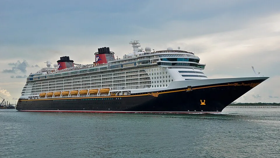 The Disney Fantasy cruise ship sailing on the ocean. The massive ship features a classic black hull, a white superstructure, yellow lifeboats, and two distinctive red funnels adorned with Mickey Mouse silhouettes.