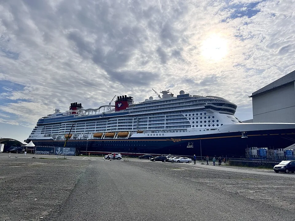 A massive Disney cruise ship named "Disney Treasure" is docked next to a shipyard under a cloudy sky with the sun shining through. The dark-hulled ship has white upper decks, yellow lifeboats, and two iconic red-and-black funnels. Cars and a few people are visible on the ground below.