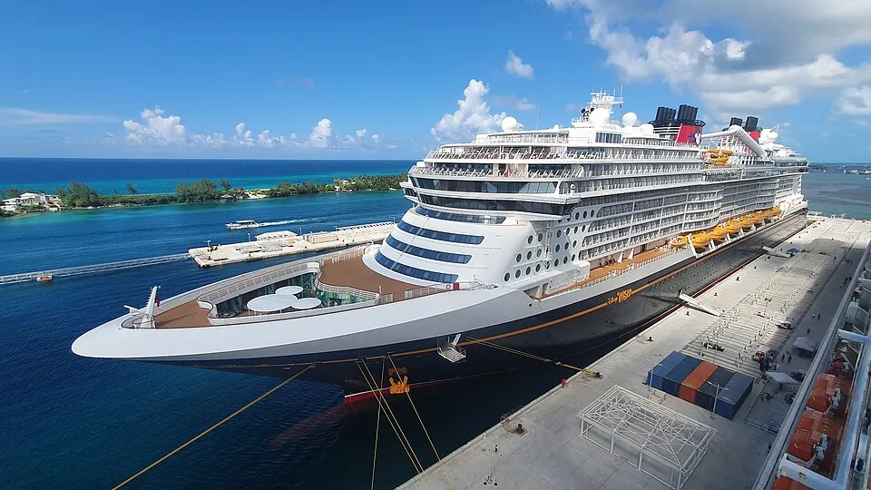An elevated view of the Disney Wish cruise ship docked at a pier next to bright blue water. The massive vessel features a classic dark hull, white decks, yellow lifeboats, and red funnels under a partly cloudy sky.