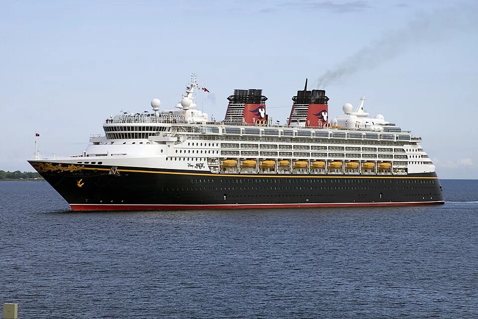 The Disney Magic cruise ship sails on calm, open blue water under a clear sky. The classic vessel features a distinctive black hull with a red stripe, a white upper body with yellow lifeboats, and two prominent red and black funnels adorned with the Mickey Mouse logo.