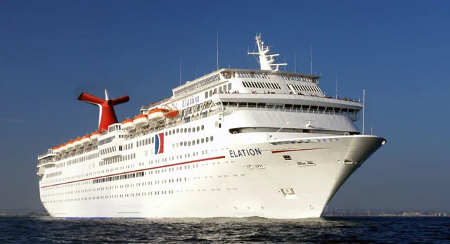 Large white Carnival Elation cruise ship sailing through calm blue water, viewed from the front side, with multiple decks, rows of windows, lifeboats, and the ship’s distinctive red-and-blue funnel against a clear sky.