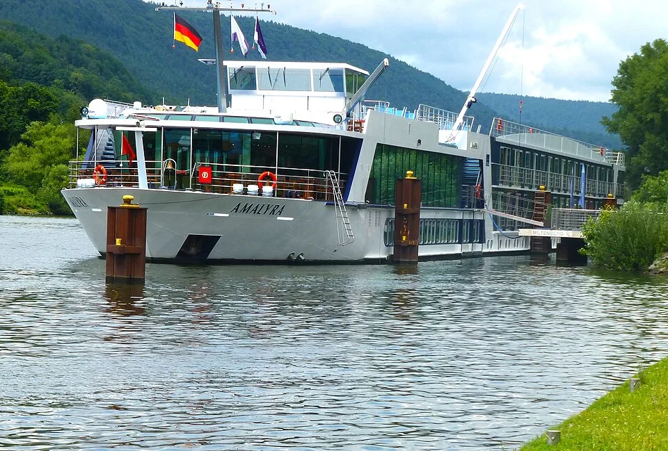 A white river cruise ship named "AmaLyra" docked at a river station. The vessel features glass windows, life rings, and several flags flying from its bow, including the German flag, set against a backdrop of dense green forested hills under a cloudy sky.