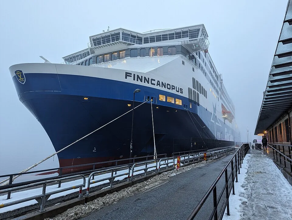 A large passenger ferry named "FINNCANOPUS" sits docked next to a snowy pier on a foggy day. The vessel has a prominent blue lower hull, a white superstructure with multiple tiers of windows, and a large bridge at the top. Metal railings run alongside the slushy concrete dock.