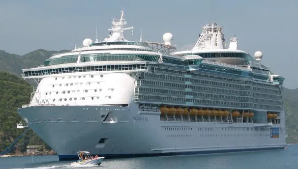 A massive white Royal Caribbean cruise ship, named "Freedom of the Seas," floating on calm blue water with a small boat in front of it. Lush green mountains are visible in the background under a hazy sky.