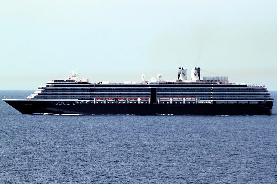 The Holland America Line cruise ship MS Oosterdam sails across open blue water. The massive vessel features a long black hull, multiple tiers of white passenger decks with orange lifeboats, and two white smokestacks near the rear against a clear sky.