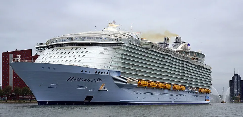 The massive white cruise ship Harmony of the Seas sails on a waterway near land with buildings under a grey sky. Yellow lifeboats are visible along the side, and a small water boat sprays a fountain in the distance.