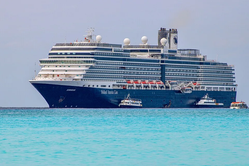 A large Holland America Line cruise ship named "EURODAM" floats on bright turquoise water under an overcast sky. The ship has a dark blue hull, white upper decks, orange lifeboats, and a white funnel with a logo. Two smaller tender boats are positioned alongside its hull.