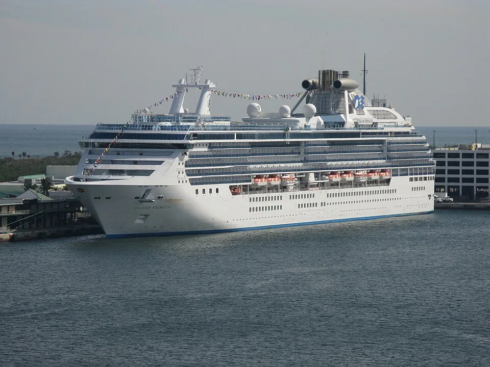 The Island Princess cruise ship is docked in calm blue water near a port. The large white vessel features rows of cabin balconies, bright orange lifeboats, and strings of colorful flags draped across its upper decks under a hazy sky.