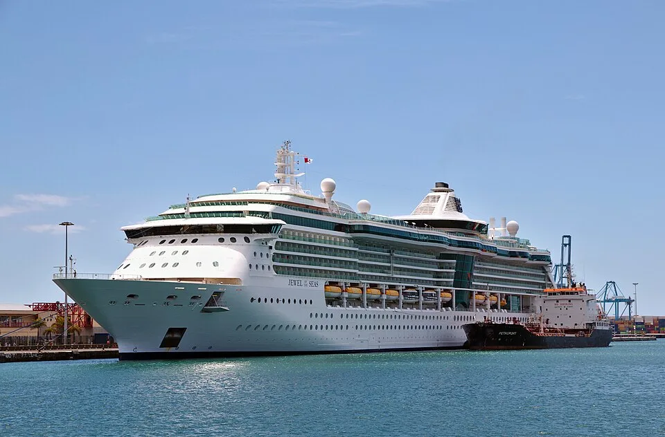 A large white Royal Caribbean cruise ship, named "Jewel of the Seas," docked in a port next to a smaller black cargo ship. The multi-story ship sits in calm blue water under a clear sky, with harbor cranes visible in the background.