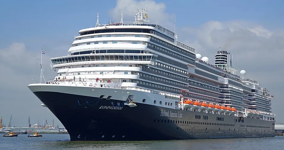The Holland America Line cruise ship Koningsdam sailing on a clear day. The large vessel has a dark blue hull, multiple white passenger decks, and a row of orange lifeboats along the side under a blue sky with light clouds.