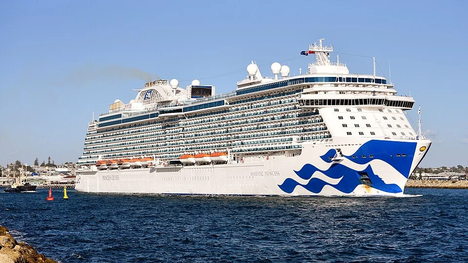 The Majestic Princess cruise ship sails on deep blue water near a coastline. The massive white vessel features vibrant blue wave designs on its bow, a multi-tiered front superstructure, thousands of cabin balconies, and a line of orange lifeboats.