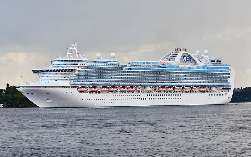The Emerald Princess cruise ship sails on open water under an overcast sky. The massive white vessel features rows of bright blue glass balconies, a line of orange lifeboats, and the distinctive Princess Cruises logo on its upper deck structure.