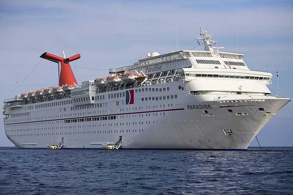 The massive Carnival Paradise cruise ship floats on deep blue water under a partly cloudy sky. The white vessel features several decks of windows, small tender boats attached near the hull, its name clearly written on the bow, and a signature red whale-tail funnel on top.