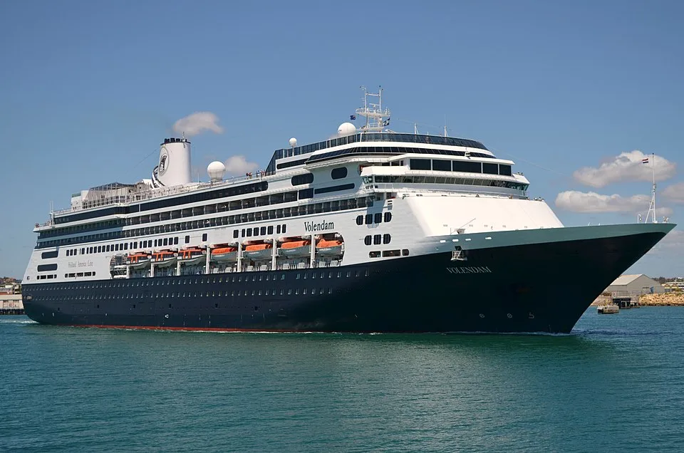 The Holland America Line cruise ship Volendam sailing on green-blue water. The large vessel has a deep blue hull, a white superstructure with multiple decks, orange lifeboats, and a white funnel under a bright blue sky.