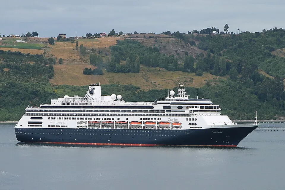 A large Holland America Line cruise ship named "Zaandam" sails on calm water. The ship features a dark blue hull with a red waterline stripe, white upper decks, orange lifeboats, and a single white funnel with a logo. In the background, a green, rolling hillside rises under an overcast sky.