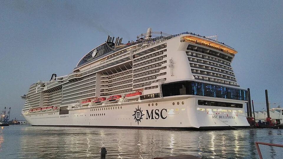 A large white cruise ship named "MSC BELLISSIMA" sits in calm water during dusk. The massive vessel features countless tiered balconies, red lifeboats, a dark glass stern window section, and a large black funnel with a star logo near the top. The MSC logo is printed on its white side.