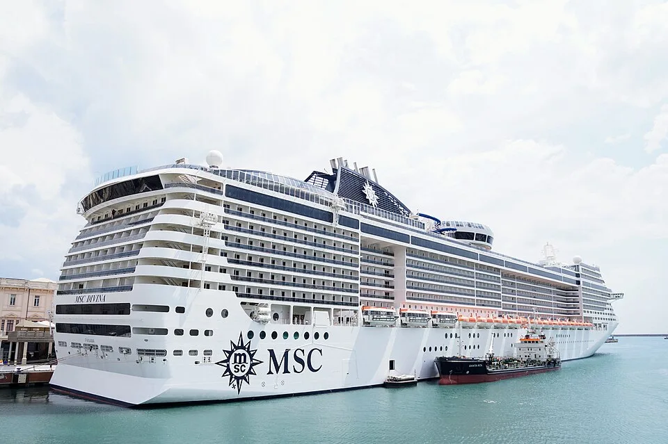 A large white MSC cruise ship, labeled "MSC Divina," is docked in calm, light blue water. A smaller red and black boat is stationed next to its side. The multi-story ship features rows of balconies and a bright, slightly overcast sky fills the background.