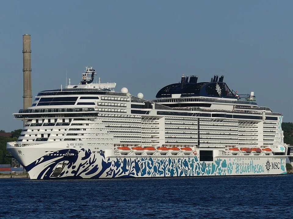 The massive cruise ship MSC Euribia floats on blue water under a clear sky. The vessel features a white hull decorated with blue marine life illustrations and the words "#savethesea," multiple tiers of passenger balconies, orange lifeboats, and a dark funnel structure.
