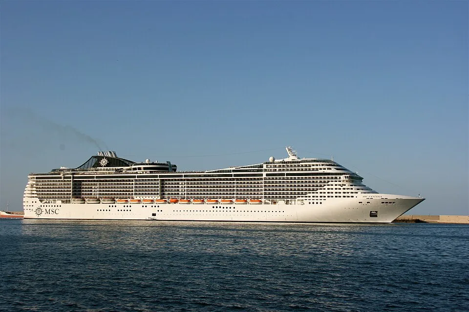 A large white cruise ship named "MSC FANTASIA" sails on deep blue water under a clear sky. The multi-deck vessel features rows of small windows, balconies, orange lifeboats along the side, and a black funnel with a star logo near the back emitting dark exhaust.