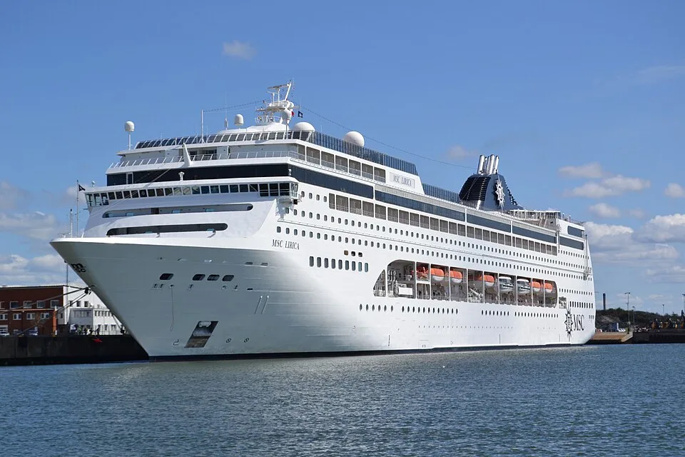 The large cruise ship MSC Lirica is docked at a port terminal on calm water. The white vessel features its name near the bow, multiple rows of square windows, orange lifeboats tucked along its side, and a dark blue funnel with the MSC logo under a bright blue sky.
