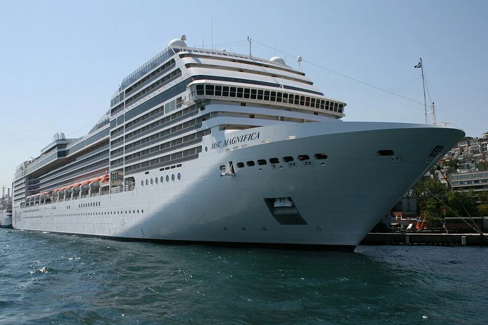 The massive cruise ship MSC Magnifica is docked in a port on choppy blue water. The white ship features multiple tiers of cabins, orange lifeboats along the side, and its name clearly written near the bow under a bright, clear sky.