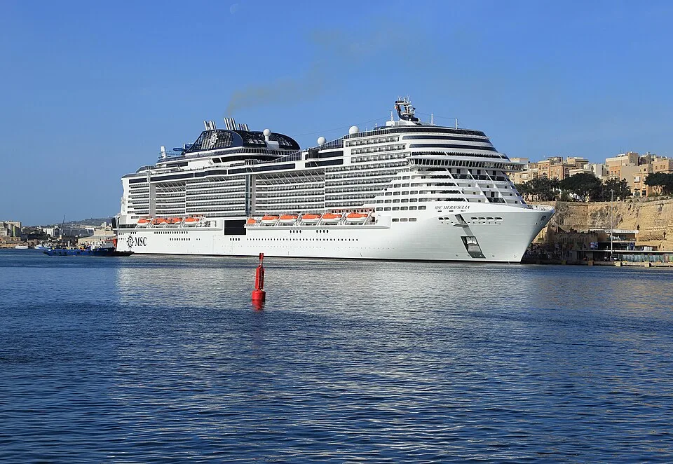 The massive cruise ship MSC Meraviglia sails through a harbor under a clear blue sky. The white vessel has a multi-tiered front, orange lifeboats, and a dark funnel emitting light smoke. A single red buoy floats in the blue water in the foreground.