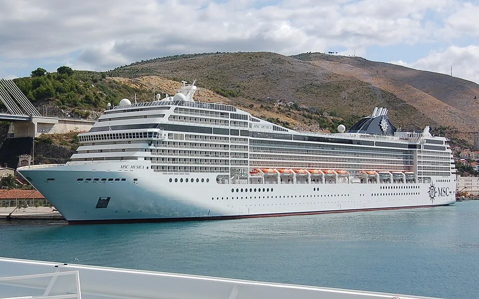 The massive cruise ship MSC Musica floats on turquoise water near a coastline. The white vessel features multiple decks with rows of balconies, orange lifeboats along the side, and a dark blue funnel with the MSC logo, set against a backdrop of arid hills and a bridge.