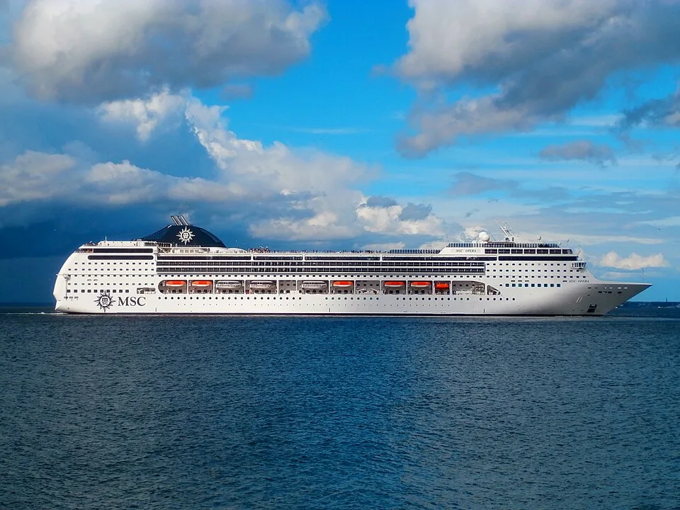 A large white cruise ship named "MSC OPERA" sails on deep blue water under a bright sky filled with large puffy clouds. The ship features many rows of windows, orange lifeboats along its side, and a large black funnel near the rear with the white MSC compass logo.