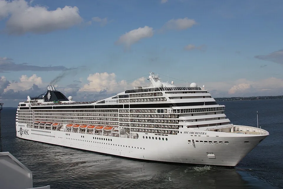 The MSC Orchestra cruise ship sails on calm open water under a blue sky with scattered clouds. The large white vessel features rows of cabin balconies, orange lifeboats along the side, and a dark funnel towards the back releasing light smoke.