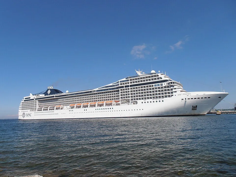The large white cruise ship MSC Poesia floats on calm ocean water under a clear, bright blue sky. The vessel features its name near the bow, multiple rows of windows and glass passenger balconies, orange lifeboats along the side, and a dark blue funnel with the MSC logo at the rear.