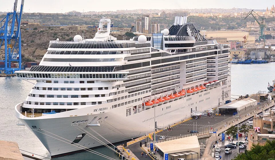A massive white cruise ship named "MSC PREZIOSA" is docked at a city port. The vessel features tiered passenger decks, rows of dark glass windows, a black funnel with a star logo, and orange lifeboats lined along the side. Port structures and a hilly city landscape are visible in the background.