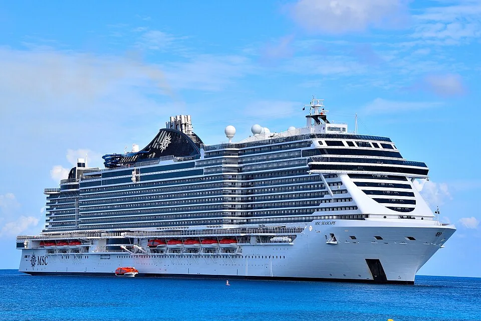 The massive cruise ship MSC Seascape sails across bright blue water under a partly cloudy sky. The white vessel features a heavily tiered superstructure with hundreds of balconies, orange lifeboats, and a large black funnel decorated with the MSC logo.