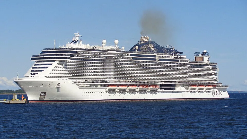 The massive cruise ship MSC Seaview sails across deep blue water under a clear sky. The white vessel features a unique condo-style design with tiered decks and hundreds of balconies. Dark smoke rises from its large black funnel toward the back.