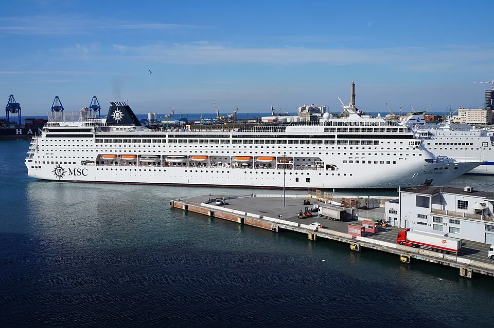 The large white cruise ship MSC Sinfonia is docked at a bustling port terminal next to a concrete pier. The ship features a dark blue funnel with the MSC logo, rows of square windows, orange lifeboats, and trucks parked on the dock under a clear blue sky.