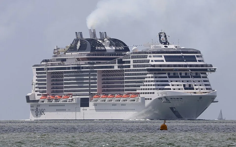 The massive cruise ship MSC Virtuosa sails on choppy water under an overcast sky. The white vessel features a highly tiered front superstructure, numerous cabin balconies, and orange lifeboats. Thick white smoke rises from its large black funnels.
