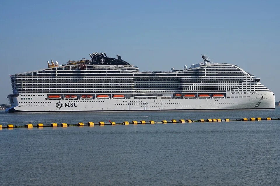 The massive cruise ship MSC World America floats on calm water under a clear blue sky. The futuristic white ship features a unique architectural design, multiple rows of balconies, orange lifeboats, and a black funnel structure. A yellow floating barrier sits in the foreground water.