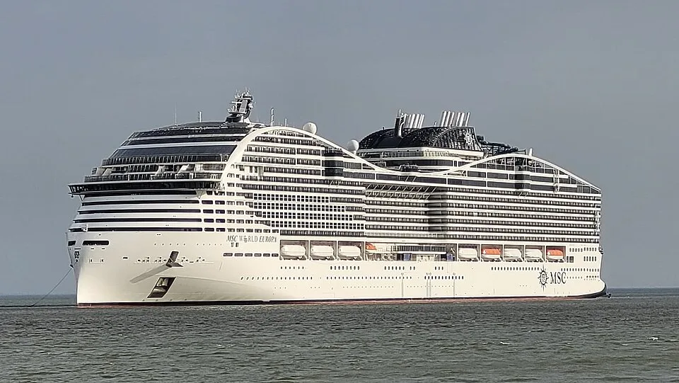 The massive cruise ship MSC World Europa floats on calm water under a hazy sky. The futuristic white vessel features a unique Y-shaped architecture at the back, multiple tiers of windows, orange lifeboats along the side, and a prominent black funnel structure.