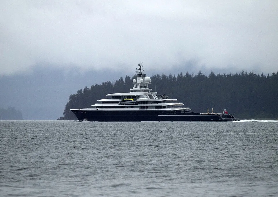A massive luxury superyacht sails on choppy gray water. The vessel features a dark hull, a multi-tiered white superstructure with open decks, and a tall radar mast. A dense green forest of pine trees lines the shore under a hazy, overcast sky.