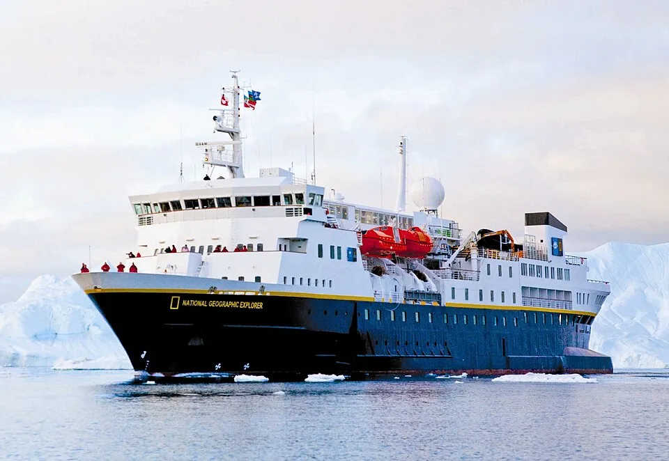 The National Geographic Explorer cruise ship sails through icy waters filled with small ice chunks. The ship features a black hull with a bright yellow stripe and a multi-level white superstructure. Passengers line the deck against a background of snowy icebergs.