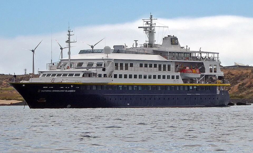 The expedition ship "NATIONAL GEOGRAPHIC ISLANDER II" floats on calm water. The multi-deck vessel features a dark blue lower hull with a yellow stripe and a white superstructure above. Wind turbines and a barren rocky shoreline are visible in the background under a cloudy sky.