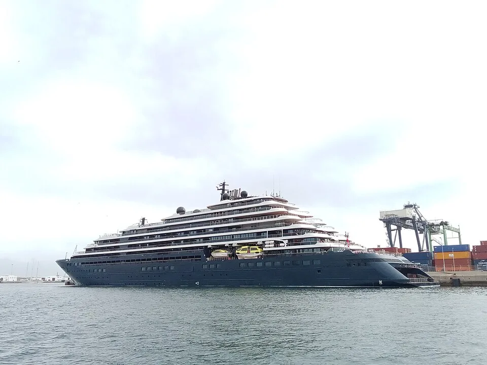 A sleek, modern luxury superyacht-style cruise ship, named "Evrima," docked at a busy port. The multi-tiered ship features a dark grey hull, tiered white decks, and yellow lifeboats, set against an overcast sky with cargo cranes visible in the background.