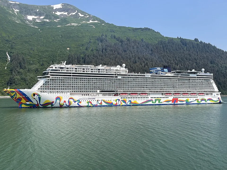 The massive cruise ship Norwegian Encore sails on calm green water. The white vessel features a vibrant, colorful abstract design swirling along its hull, multiple tiers of balconies, orange lifeboats, and blue water slides on top, set against a backdrop of a lush green mountain.