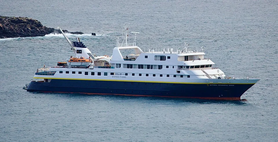 An elevated shot of a National Geographic cruise ship on the water. The ship features a dark blue lower hull with a yellow stripe, multiple white upper decks, and a prominent white arch. The text "EXPEDITIONS" is visible on the side. A small rocky island with breaking waves is in the upper left.