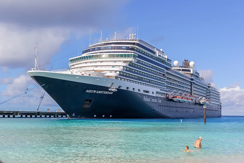 The large cruise ship MS Nieuw Amsterdam from Holland America Line is docked at a pier in clear turquoise water. Two people swim in the foreground shallows under a blue sky with fluffy clouds.